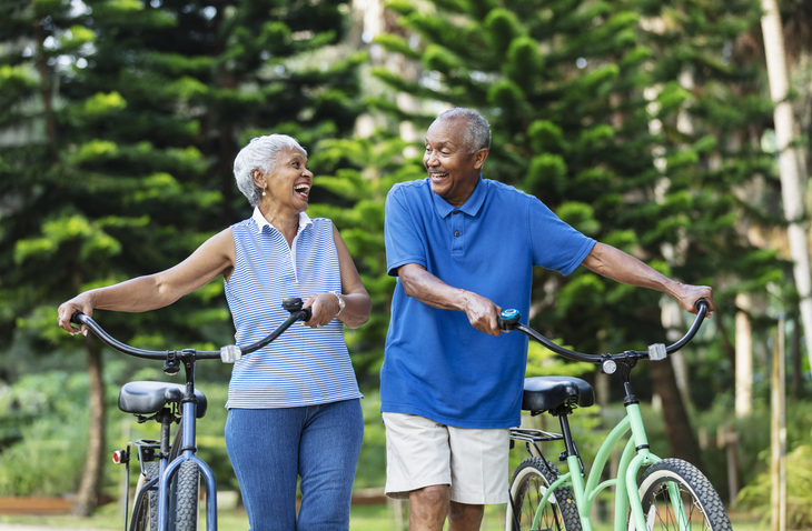 A senior African American couple in the park, walking side by side with their bicycles, looking at each other. The man is smiling and the woman is laughing.